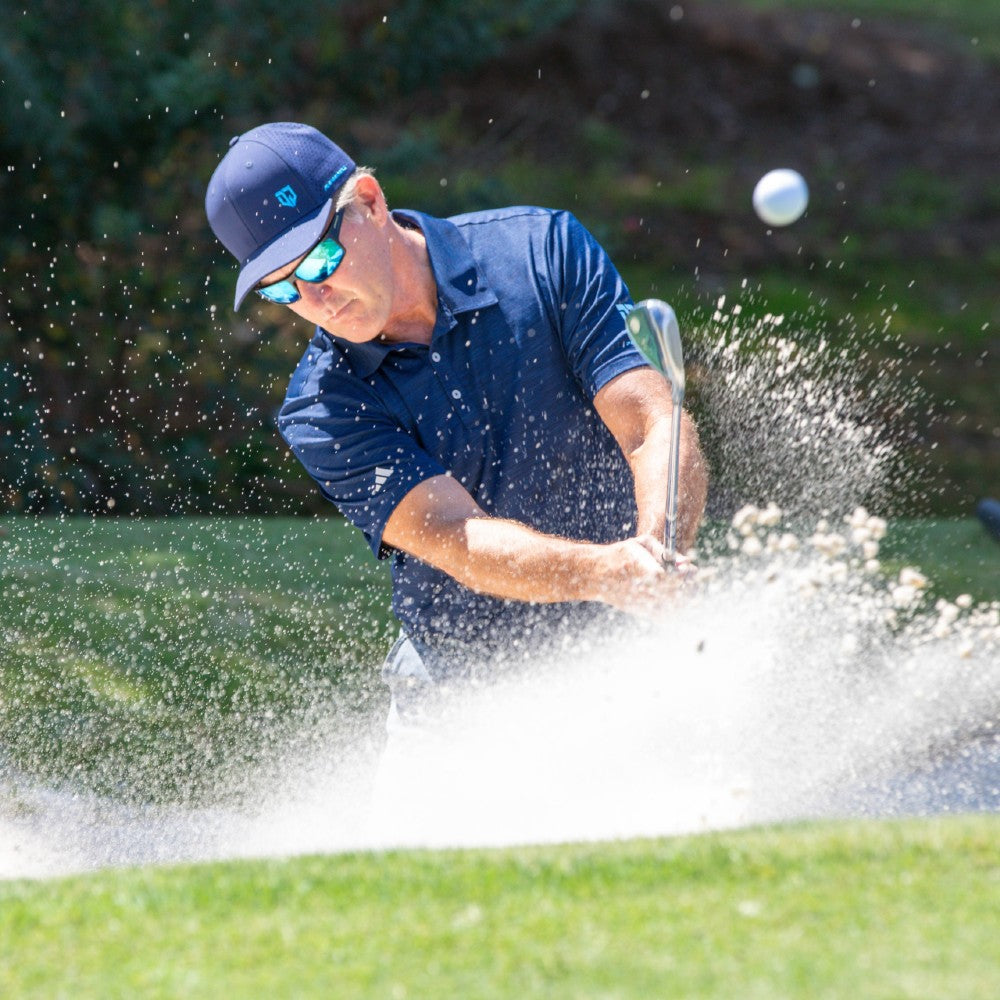 Dan Jansen hitting a golf shot from the bunker with sand flying, wearing Jansen Eyewear
