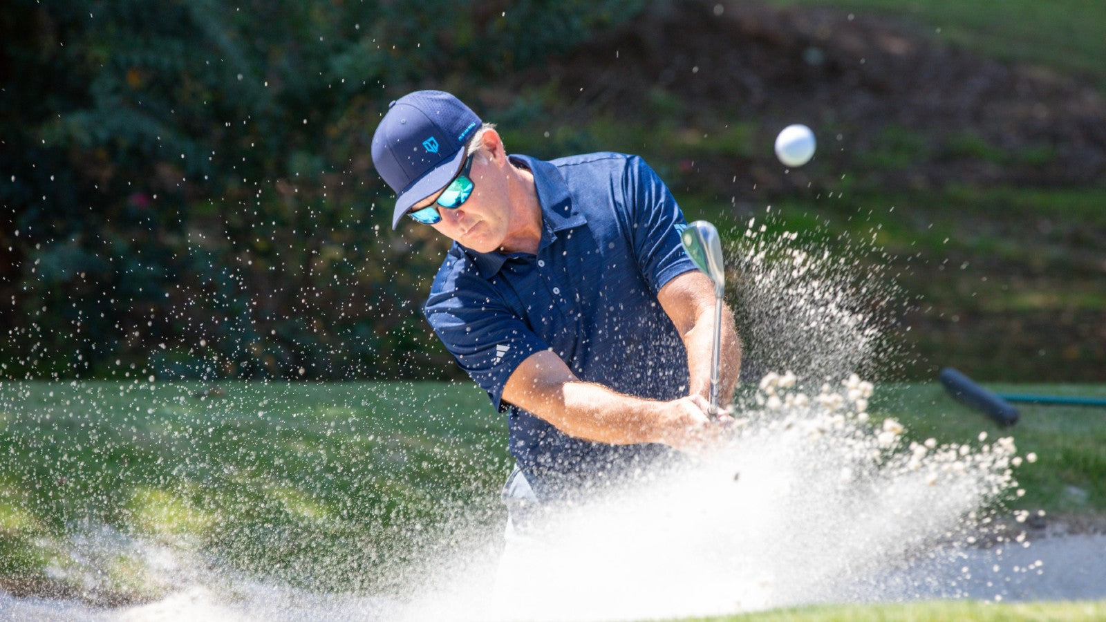 Dan Jansen hitting a bunker shot with sand flying, wearing Jansen Eyewear for focus and clarity